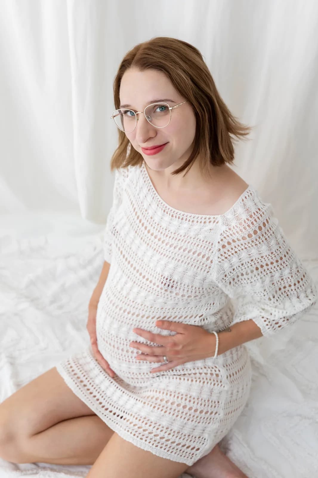 Těhotenské focení v ateliéru, maminka sedí na bílé posteli v háčkovaných šatech a drží si bříško