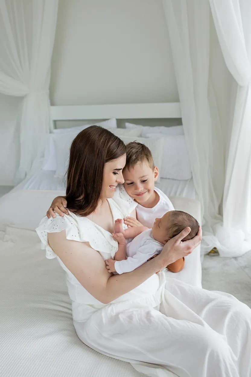 Náhled průvodce newborn focením, maminka v bílých šatech drží novorozenou holčičku, vedle starší syn, bílá ložnice ve skandinávském stylu, Marcela Kramářová, Fotoatelier Brno